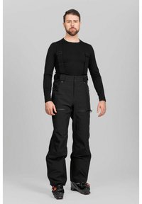 Mies seisoo harmaalla taustalla pukeutuneena mustaan pitkähihainen paitaan, mustiin lumihousuihin, joissa on henkselit, ja mustiin suksikenkiin.