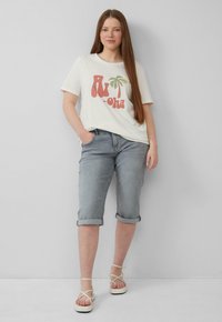 Lichtgewicht crème T-shirt met retro 'Aloha' opdruk, gecombineerd met opgerolde lichtgrijze denim shorts en beige sandalen met bandjes.