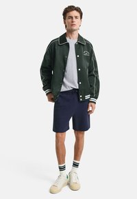 Groene varsityjack met witte bies, grijs T-shirt, marineblauwe shorts en witte sneakers met groene accenten en gestreepte sokken.