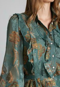 Blusa floral verde con acentos de volantes, con patrones bordados en oro y óxido. Fabricada en un tejido ligero y translúcido con botones.