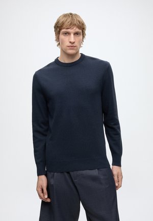 Young man with blond hair wearing a dark navy blue crewneck sweater and matching loose trousers, standing against a plain light background.