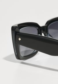 Óculos de sol grandes e pretos com lentes escuras. As hastes têm um acabamento brilhante e um pequeno detalhe de logótipo na lateral.