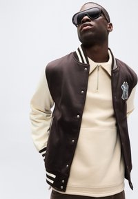 Άνδρας φορώντας υπερμεγέθη μαύρα γυαλιά ηλίου, κρεμ ζιπ-επ τοπ και καφέ αθλητικό μπουφάν με άσπρα μανίκια και λογότυπο NY, ποζάρει μπροστά σε απλό φόντο.