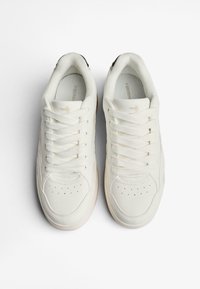 Biele tenisky s hladkým povrchom, okrúlou špičkou a perforovanými detailmi. Obsahujú šnúrky, textúrovaný jazyk a gumenú podrážku.