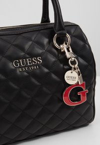 Sac à main noir matelassé avec fermeture éclair dorée, logo GUESS en métal et charme attaché avec étiquette "Guess Est. 1981" et lettre rouge G.