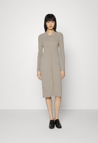 Vestido de malha em bege claro, com mangas longas, colarinho e fecho frontal com botões. Textura suave, forma ajustada e design até aos joelhos. Sapatos pretos.