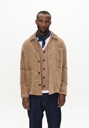 Chaqueta de pana marrón con cuello, bolsillos frontales y cierre de botones, superpuesta a un cárdigan marrón y una camisa blanca. Pantalones de mezclilla azul oscuro.