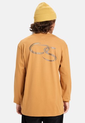 Pitkähihaisen paidan sinappikeltainen puuvilla, jossa on suuri metallinhohtoinen harmaa logo selässä, rento malli ja pyöreä kaula-aukko.