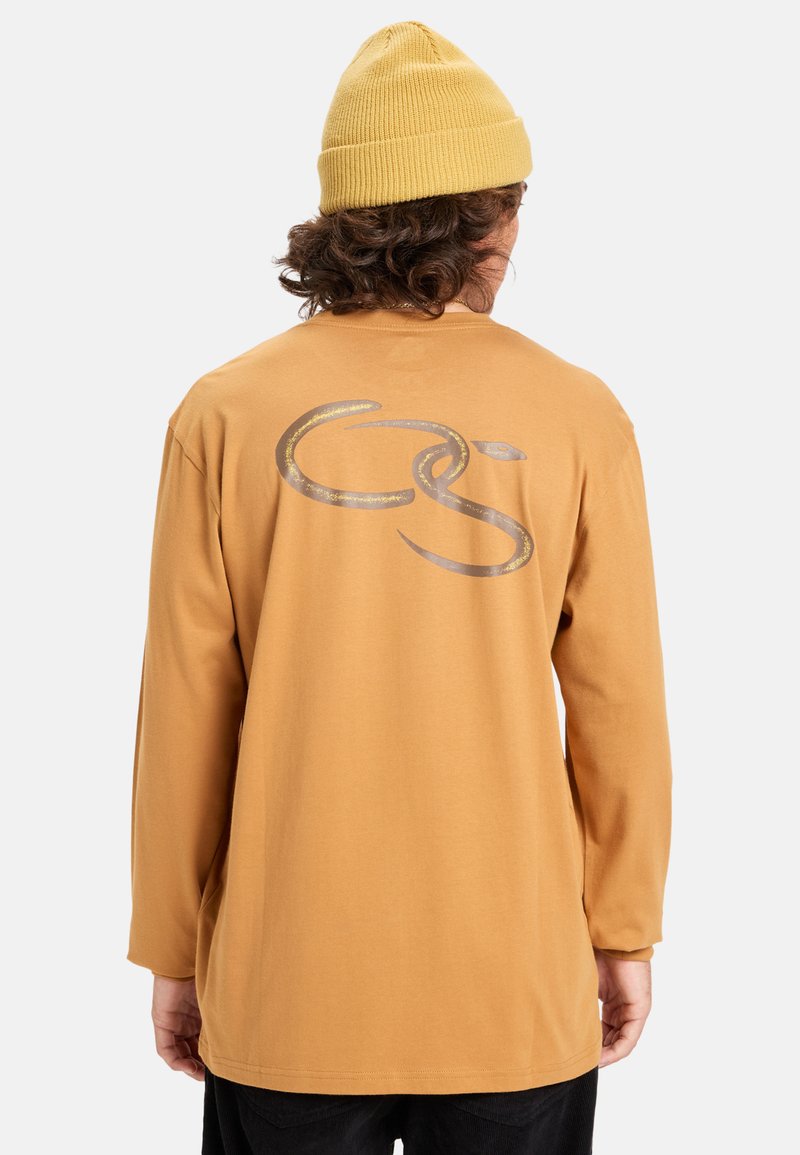 Pitkähihaisen paidan sinappikeltainen puuvilla, jossa on suuri metallinhohtoinen harmaa logo selässä, rento malli ja pyöreä kaula-aukko.