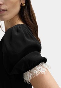 Schwarzes Oberteil mit Puffärmeln aus strukturiertem Stoff und weißem Spitzenbesatz am Bündchen. Seitenansicht mit Haaren des Models, die herabfallen. Goldene Hula-Hoops.