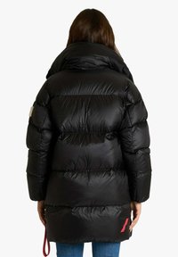 Giacca a vento nera con collo alto, texture trapuntata e dettaglio rosso all'orlo. Vista posteriore che mostra una vestibilità ampia con una lunghezza leggermente maggiore.