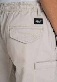 Pantalones cortos beige de cargo con cintura elástica, que cuentan con un bolsillo trasero con tapa y una etiqueta negra cosida en la cintura.