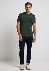 Camiseta de algodón verde con mangas cortas, que presenta un diseño gráfico negro de montañas. Combinada con jeans oscuros y zapatillas blancas.