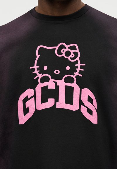 Άτομο που φοράει μαύρο μπλουζάκι με ροζ καρτούν γάτα με φιόγκο και μεγάλα ροζ γράμματα "GCDS" κάτω από τη γάτα.