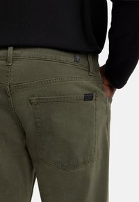 Jeans in denim verde oliva con una texture liscia, dotati di una tasca posteriore e un'etichetta con branding minimale. Design classico a cinque tasche.