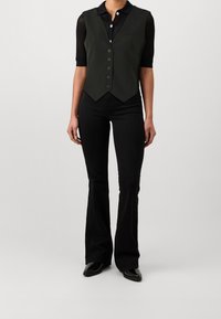 Svarta utsvängda jeans kombinerat med en olivgrön väst med spetsig nederkant, fem knappar och en bröstficka, bärs över en svart skjorta med krage.