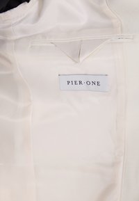 Cremfarbener Satinstoff mit glatter Oberfläche. Mit einem aufgenähten Etikett "PIER • ONE" in Schwarz und einer kleinen Tasche über dem Etikett.