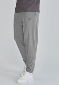 Szürke, tapered fazonú futónadrág, rugalmas mandzsettákkal. Oldalsó zsebekkel rendelkezik, a lábon fekete "SIKSILK" logóval, fehér sportcipővel párosítva.