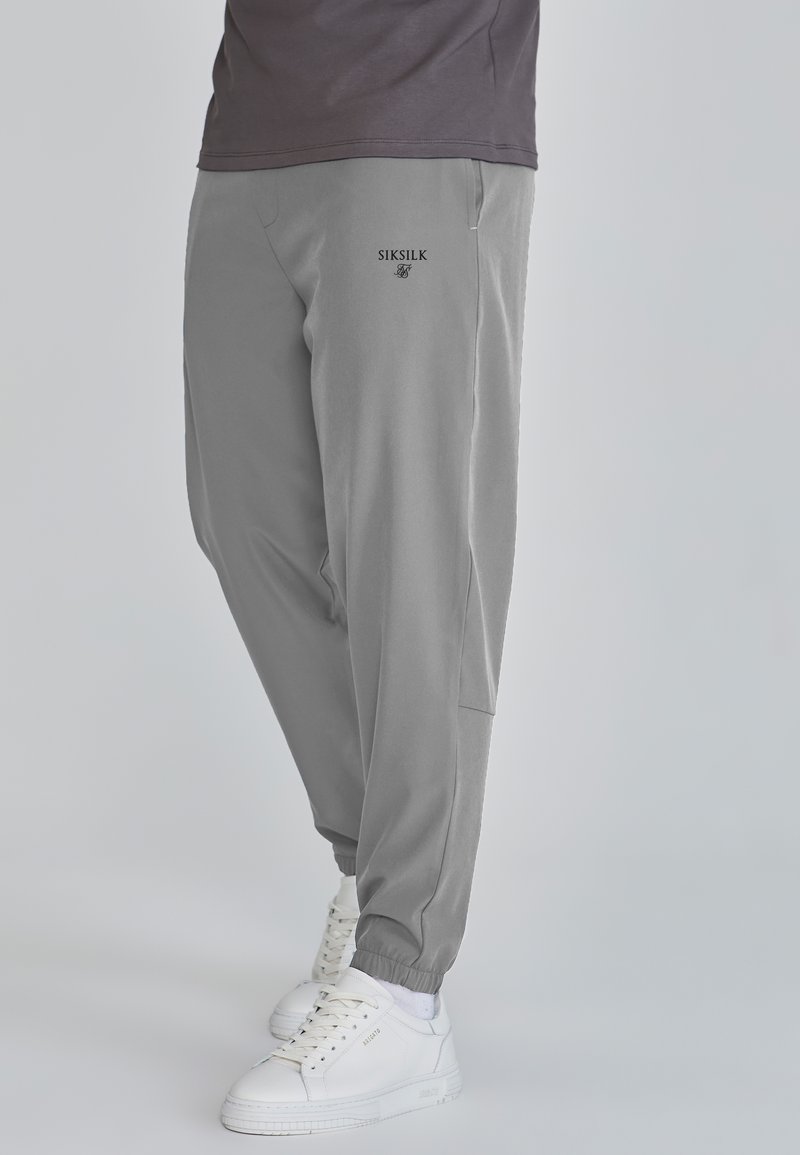 Szürke, tapered fazonú futónadrág, rugalmas mandzsettákkal. Oldalsó zsebekkel rendelkezik, a lábon fekete "SIKSILK" logóval, fehér sportcipővel párosítva.