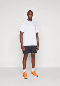 Camiseta blanca de manga corta con logo negro, combinada con pantalones cortos negros con tres rayas y zapatos deportivos naranjas.