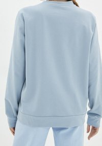 Persona vistiendo una blusa de manga larga azul clara y pantalones a juego, vista desde atrás sobre un fondo blanco liso.