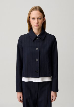 Giovane donna con capelli biondi lisci che indossa una giacca blu scuro con bottoni sopra una maglietta bianca contro uno sfondo chiaro e semplice.