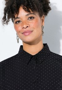 Camicia nera con colletto abbottonato, caratterizzata da pois bianchi e una texture morbida. Modello con orecchini di grande impatto e capelli ricci in vista.