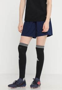 Camiseta deportiva negra, pantalones cortos azul marino, calcetas negras hasta la rodilla con rayas blancas y botas de fútbol azules con acentos rojos.