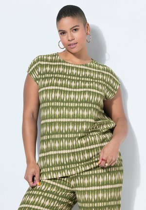 Kvinde med kort hår iført øreringe i form af ringe og en grøn mønstret kortærmet top samt matchende bukser, poserer foran en hvid baggrund.