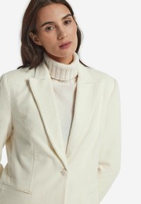 Elfenbeinfarbener gerippter Blazer mit Revers, einreihigem Knopfverschluss und seitlichen Taschen, über einem cremefarbenen Rollkragenpullover getragen. Weiche Textur.