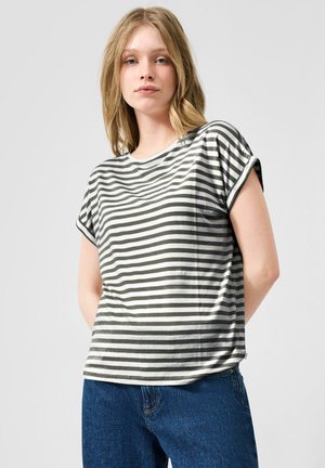 Junge Frau steht mit den Händen hinter dem Rücken, trägt ein weißes und dunkelgraues, horizontal gestreiftes Kurzarmshirt und blaue Jeans.