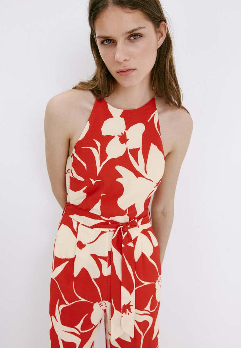 Jeune femme portant une combinaison rouge sans manches avec un grand motif floral blanc et une ceinture nouée à la taille, debout sur fond blanc.