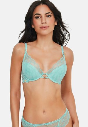 Vrouw die een lichtblauwe kanten beha en bijpassende kanten slip draagt, staande tegen een effen witte achtergrond.