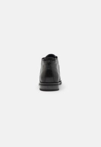 Bota de cuero negro hasta el tobillo con punta redondeada, textura suave, costuras visibles y un tacón bajo. La vista trasera muestra un diseño minimalista.