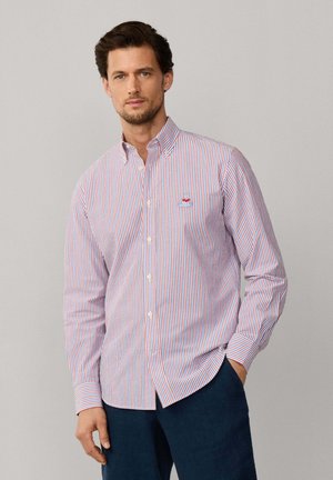 Hombre con camisa de botones a rayas verticales en rojo, blanco y azul con logo bordado, combinada con pantalones azul oscuro, de pie frente a un fondo liso.