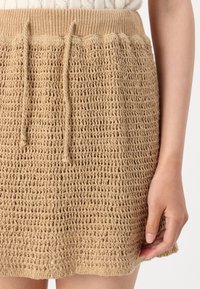 Strikket beige skjørt med snøring i livet, som har et teksturert, vevet mønster. Hempen er bølgete, og materialet ser mykt og fleksibelt ut.