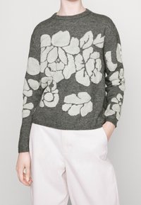 Pull en maille grise avec de grands motifs floraux blancs, porté avec un pantalon rose clair, une main dans la poche, sur un fond uni.