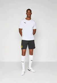 Vīriešu sportists baltā Puma krekla, melnu šortu ar dzeltenu Borussia Dortmund logotipu, baltu zeķu un baltu apavu tērpā, stāv pret vienkāršu fonu.