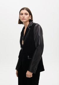 Blazer negro con solapa de satén, con un diseño estructurado y tejido de terciopelo texturizado. Incluye dos bolsillos frontales y detalles de botones.