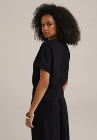 Čierny, štruktúrovaný top s krátkymi rukávmi, voľným strihom a z gathered pásom. Model má na jednom zápästí zlatý náramok.