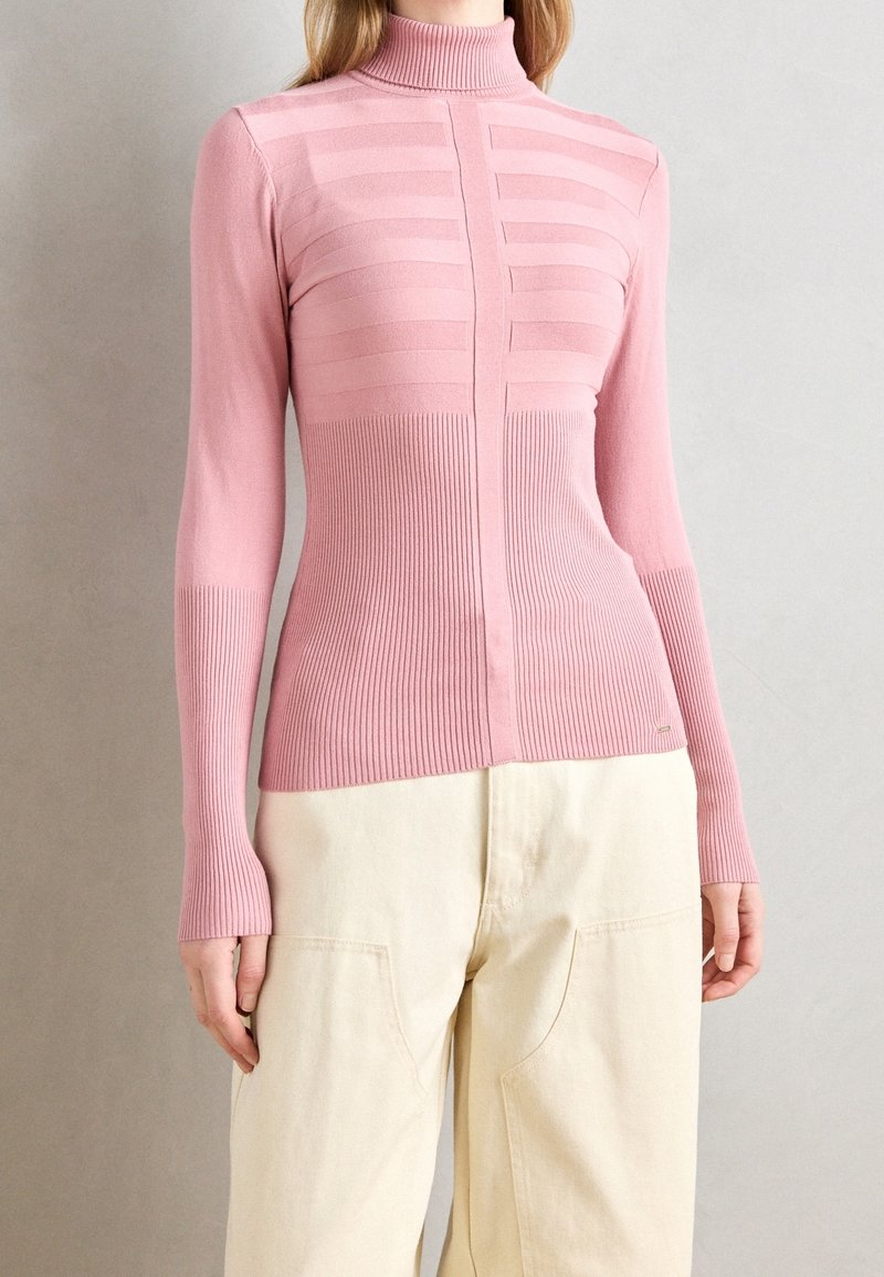 Femme portant un pull à col roulé côtelé rose ajusté avec des rayures horizontales texturées, associé à un pantalon crème taille haute.