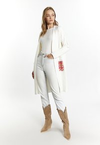 Cardigan bianco a maglia con fessure laterali, abbinato a jeans azzurri a vita alta e stivali cowboy in suede beige. Dettaglio della tasca a fantasia rosa.