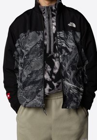 Svart och grå jacka med abstrakt meshmönster och fleecefoder, försedd med hög krage och dragkedjestängning. Detaljer inkluderar logotyp.