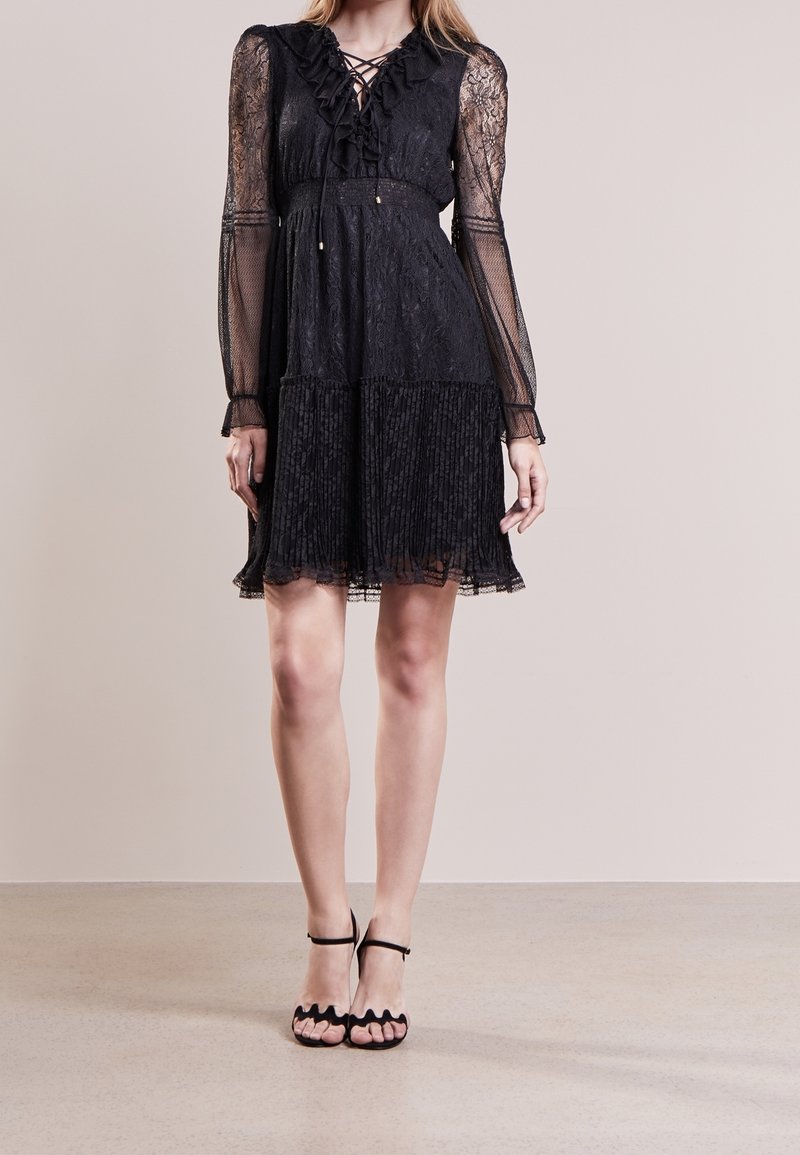 Femme portant une robe noire en dentelle avec des manches longues transparentes, des détails à volants, une jupe plissée et des talons noirs à brides festonnées, debout sur un sol beige.