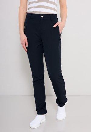 Persoon draagt zwarte nauwsluitende broek met ritssluitingen, een beige gestreept shirt en witte sneakers, staand tegen een lichtgrijze achtergrond.