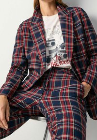 Blazer e pantaloni a scacchi in blu navy, rosso e bianco. Il blazer presenta un colletto a punta, mentre la maglietta sottostante ha una stampa grafica.