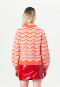 Suéter de malha com padrões ondulados em laranja e rosa, decote redondo e mangas bufantes. Combinado com uma saia mini vermelha brilhante.