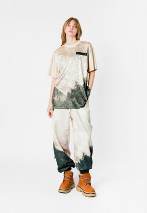 Camisa de terciopelo de manga corta en crema con un estampado de paisaje oscuro; pantalones sueltos y estampados; botas marrones con cordones y suelas texturizadas.