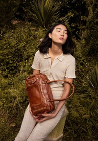 Brown leather handbag with dual handles, multiple zippered pockets, and a smooth texture. Model wearing a light beige outfit is seated.