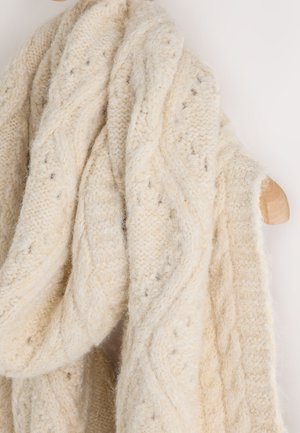 Šal krem boje, pleten grubom tehnikom s uzorkom pletenica i tekstura, prebačen preko drvenog vješalice na svijetloj pozadini.